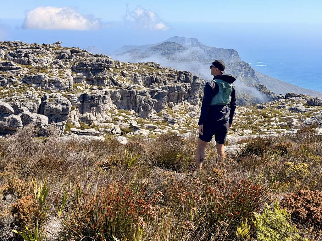 Man standing on Table Mountain looking over rocky cliffs and the Atlantic Ocean with blue sky and clouds in the distance.