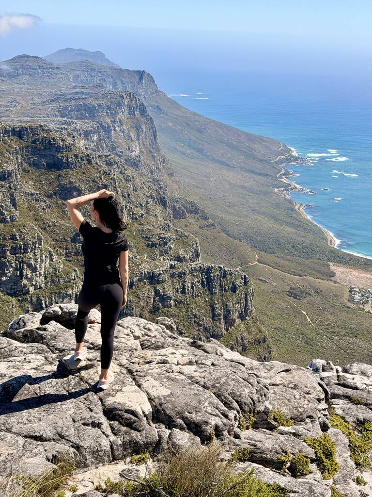 Woman standing on a rocky edge on Table Mountain overlooking the Cape Town coastline and blue ocean on a sunny day.