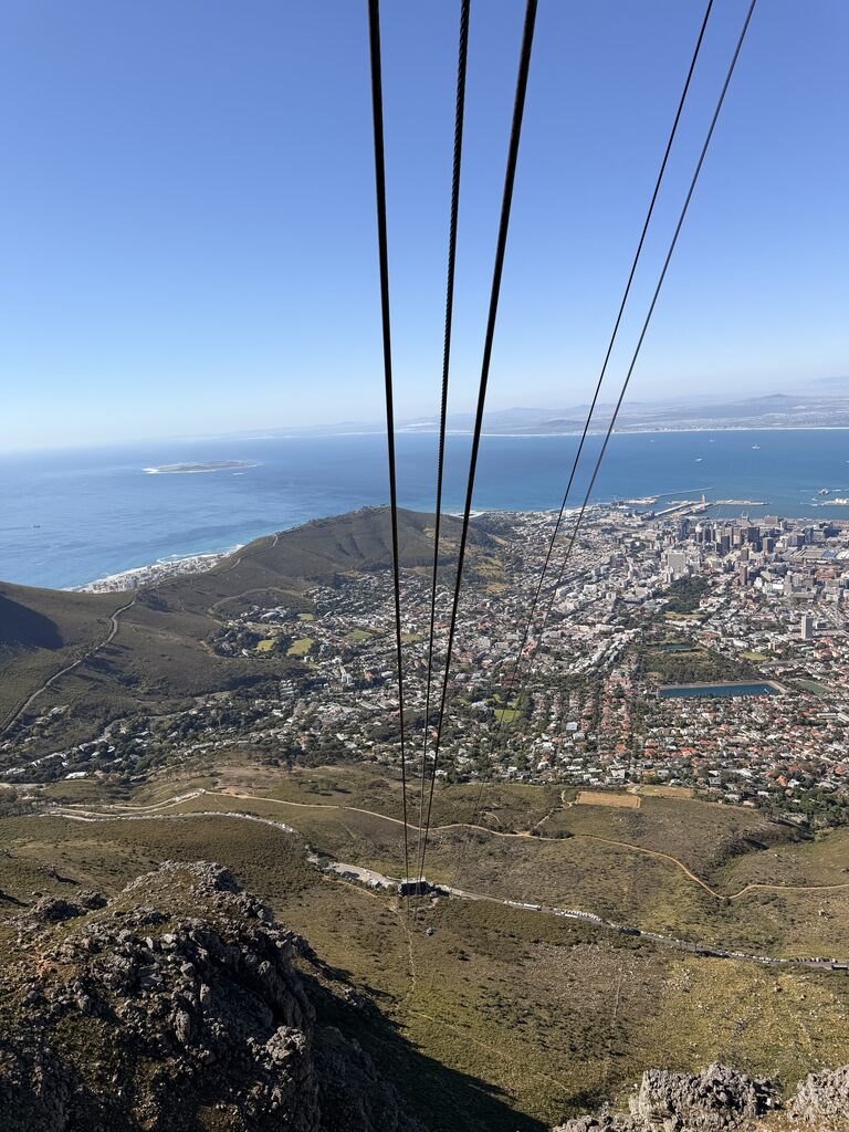 View from Table Mountain cable car looking down toward Cape Town city, coastline, and blue ocean on a clear day.
